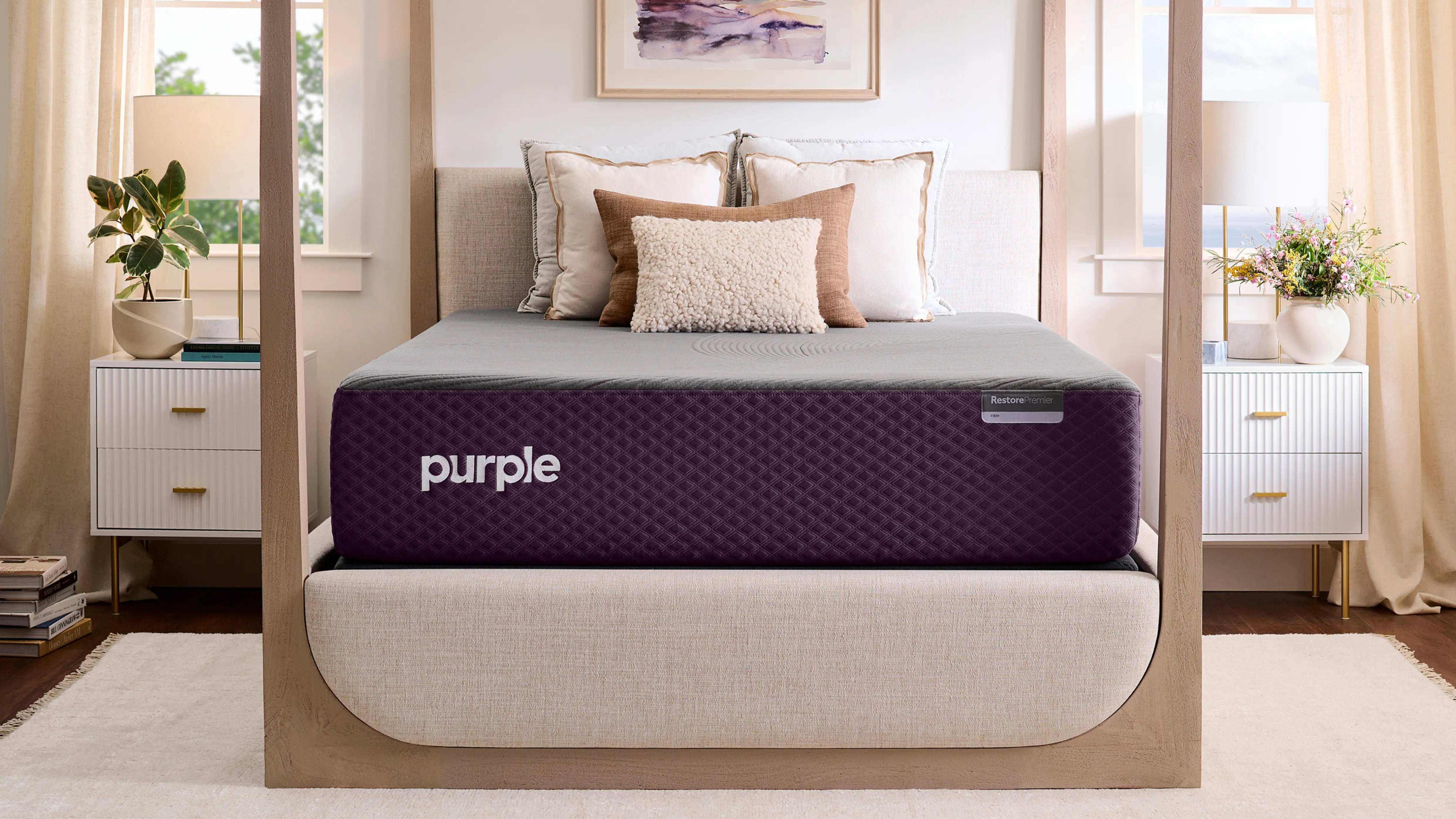A Purple brand mattress on a light-toned, four-poster bed in a bright bedroom, with neutral furniture and decorative pillows.