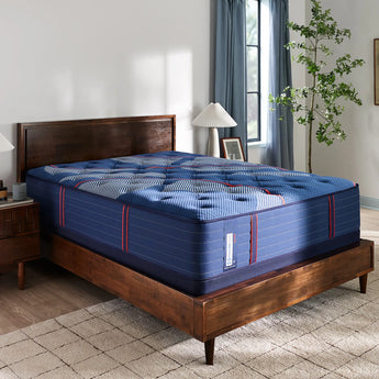 Angled view of the Sealy Posturepedic® Elite Albany 14-inch Firm Mattress, featuring a plush, quilted blue surface with red and white accents, set atop a modern wooden bed frame in a cozy, naturally lit bedroom with soft curtains and green indoor plants