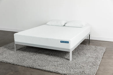 Angled view of a white Molecule mattress featuring a subtle hexagonal textured pattern, resting on a minimalist gray metal frame. The mattress is accompanied by two matching pillows and sits atop a plush gray shag rug in a modern, clean bedroom environment.