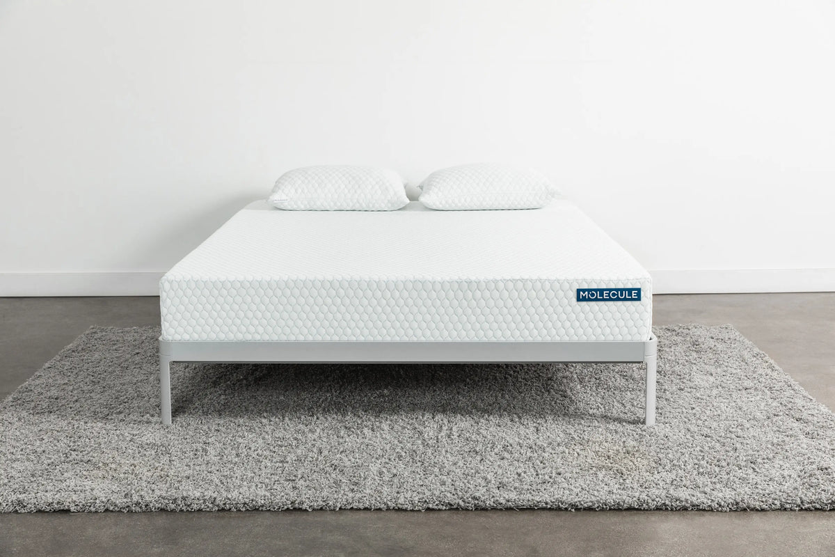 Front view of a white Molecule mattress with a textured hexagonal pattern, placed on a minimalist gray metal frame with two matching pillows on top, sitting on a gray shag rug in a clean, modern bedroom setting