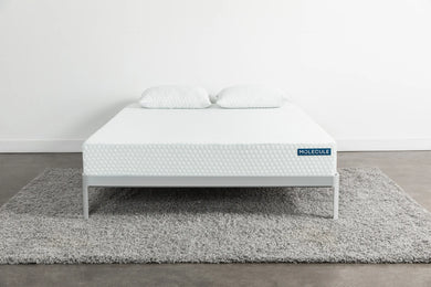 Front view of a white Molecule mattress with a textured hexagonal pattern, placed on a minimalist gray metal frame with two matching pillows on top, sitting on a gray shag rug in a clean, modern bedroom setting