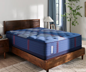 Angled view of the Sealy Posturepedic® Elite Albany 14-inch Firm Mattress, featuring a plush, quilted blue surface with red and white accents, set atop a modern wooden bed frame in a cozy, naturally lit bedroom with soft curtains and green indoor plants