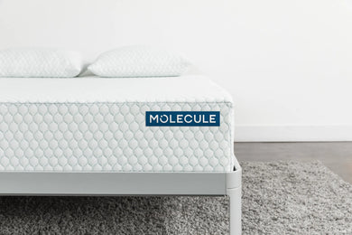 Close-up side view of the Molecule mattress highlighting the detailed hexagonal textured pattern and prominent blue Molecule branding label. The mattress sits on a sleek gray metal frame and is topped with two matching white pillows, situated in a modern, minimalist bedroom setting.
