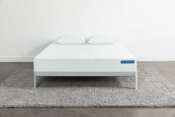 Front view of a white Molecule mattress with a textured hexagonal pattern, placed on a minimalist gray metal frame with two matching pillows on top, sitting on a gray shag rug in a clean, modern bedroom setting