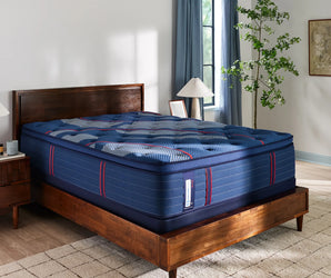 Angled view of the Sealy Posturepedic® Elite Albany 15-inch Soft Euro Top Mattress placed on a contemporary wooden bed frame in a serene, naturally-lit bedroom setting. The mattress features a plush blue quilted surface accented with stylish red and white details, conveying softness and comfort.